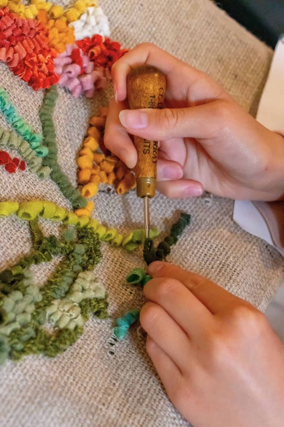 A person uses a wooden-handled punch needle to create a colorful yarn design on burlap fabric.