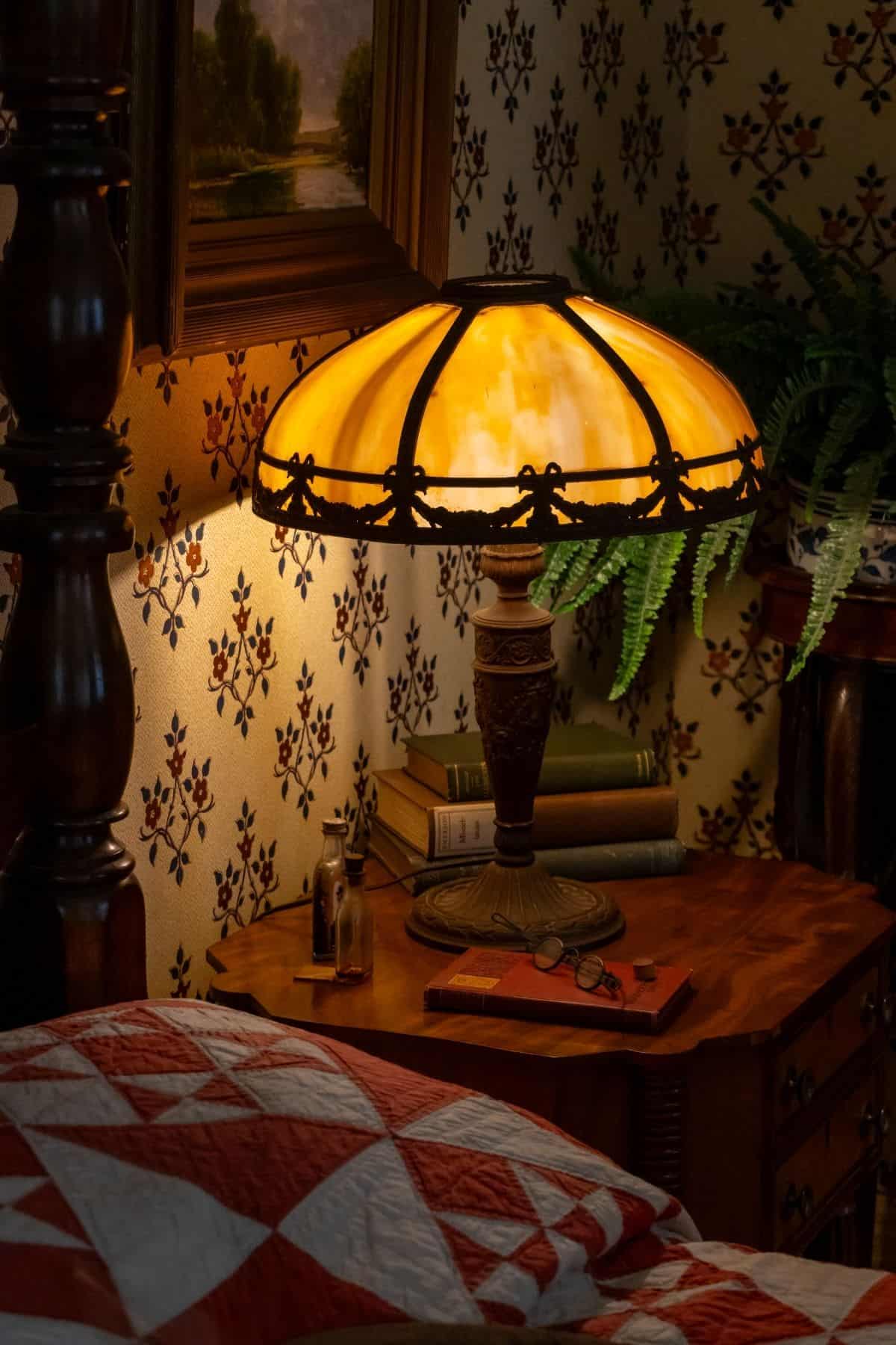 A vintage lamp with a stained glass shade sits on a wooden bedside table with books, glasses, a small figurine, and a fern, near a patterned wallpaper and a red-and-white quilt.