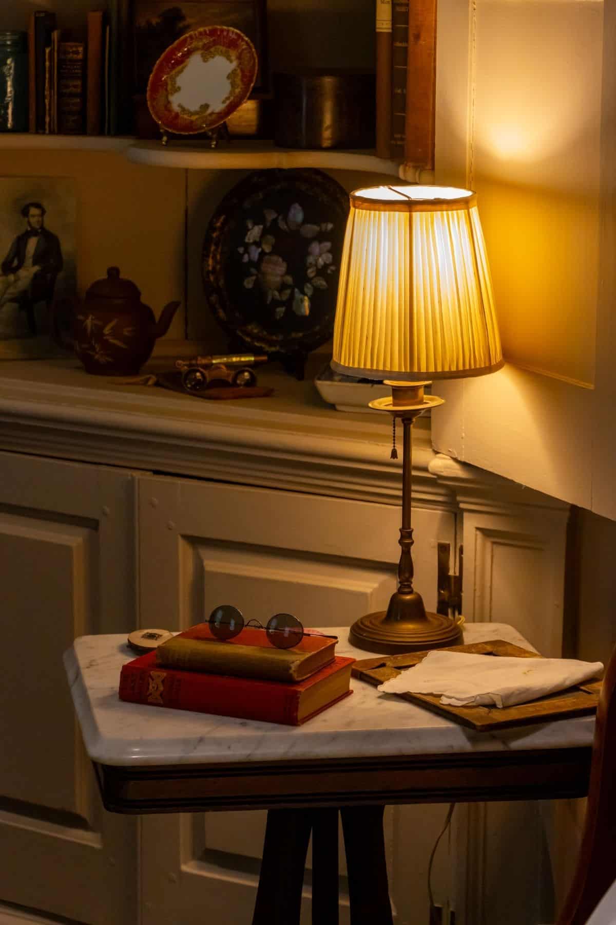 A small table with books, eyeglasses, and a lamp sits beside shelves with teapots, plates, and framed art in a warmly lit room.