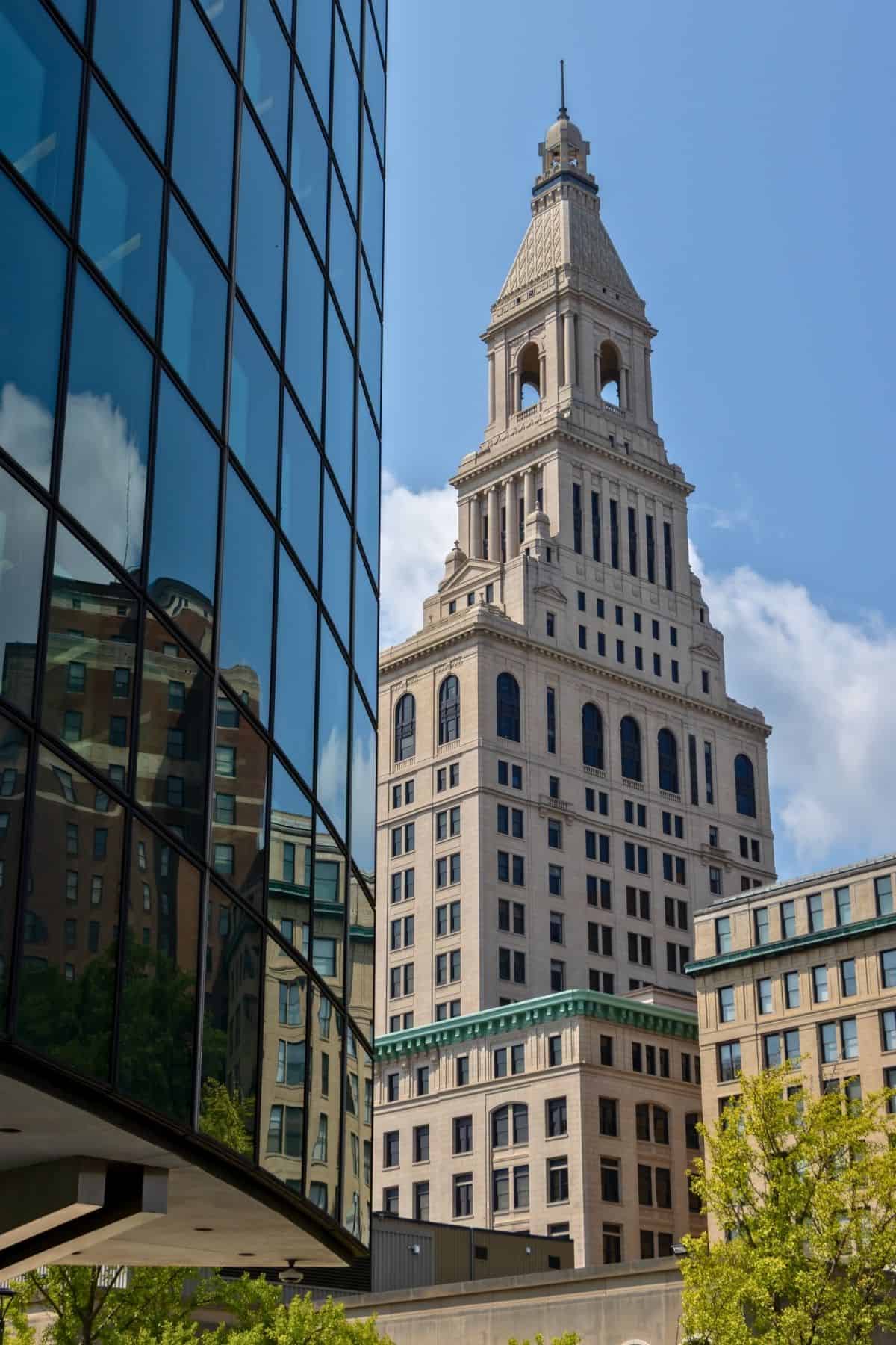 A historic stone skyscraper with a clock tower rises behind a modern glass building reflecting surrounding architecture under a clear blue sky.