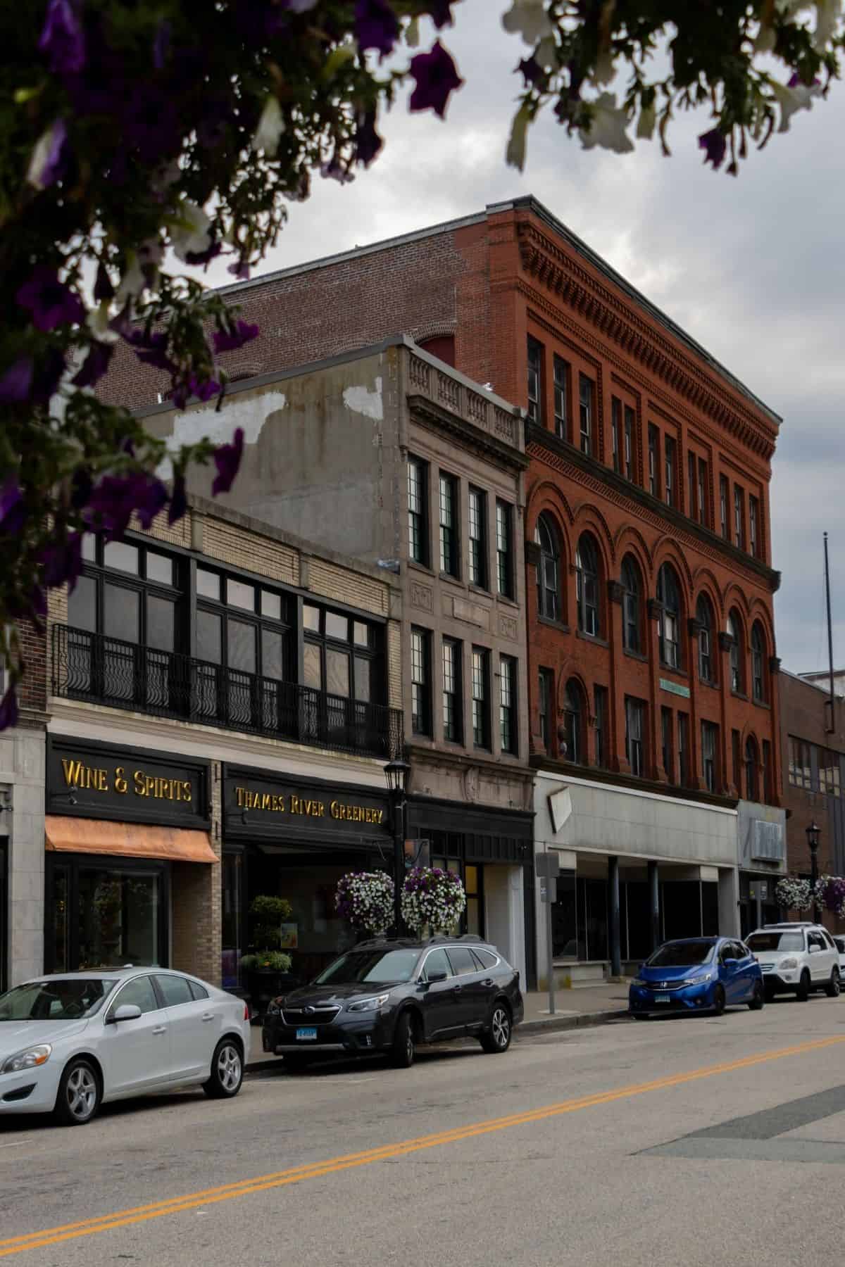 Street view of parked cars in front of brick and stone buildings, including shops with signs for “Wine & Spirits” and “Thames River Gallery” on a cloudy day.