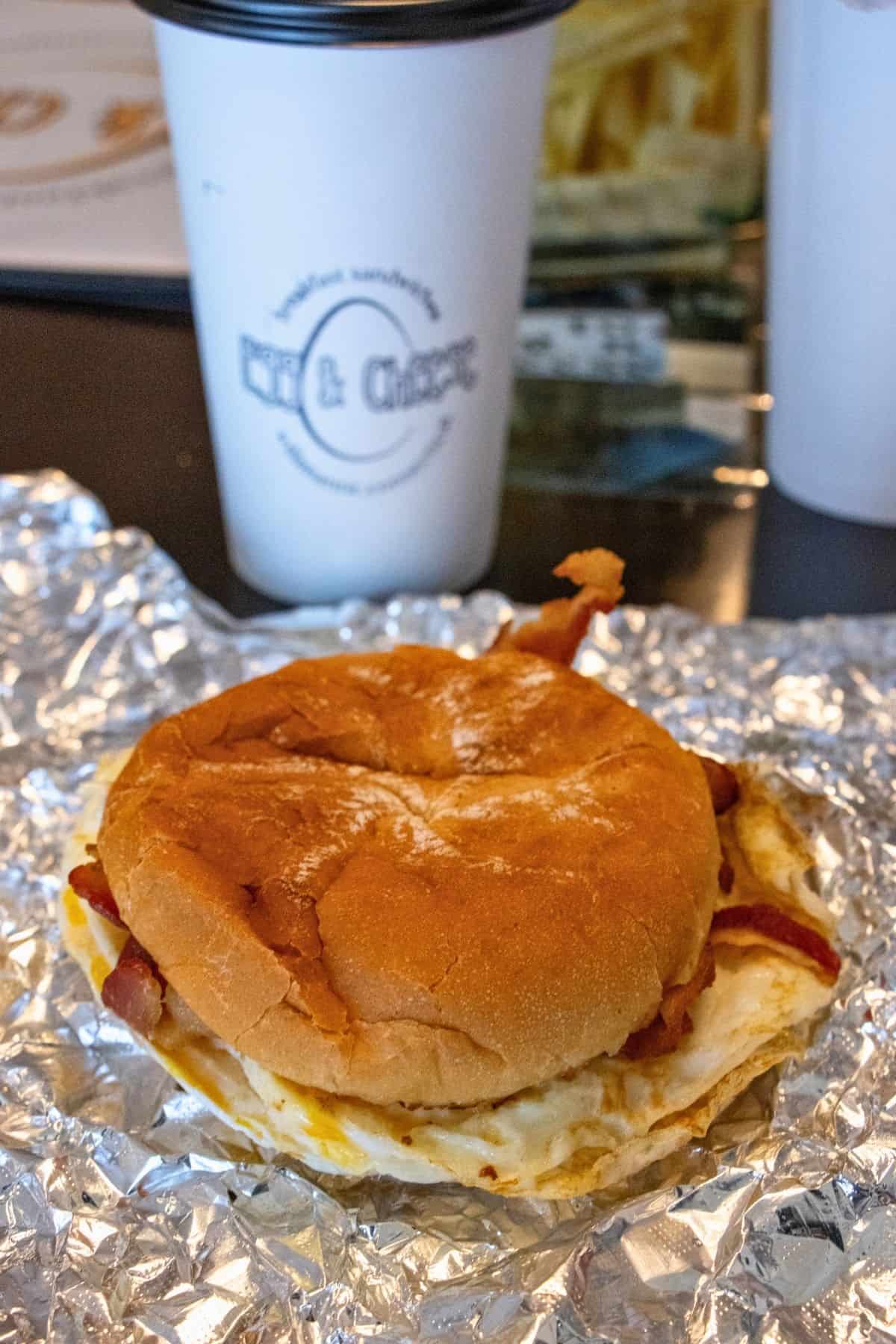 A breakfast sandwich with egg and bacon on a bun sits on foil, with a paper coffee cup and fries visible in the background.