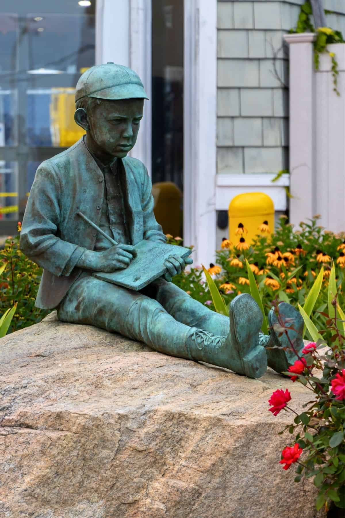 Bronze statue of a young boy in a cap sitting on a rock, writing in a notebook, surrounded by flowers and near a building.