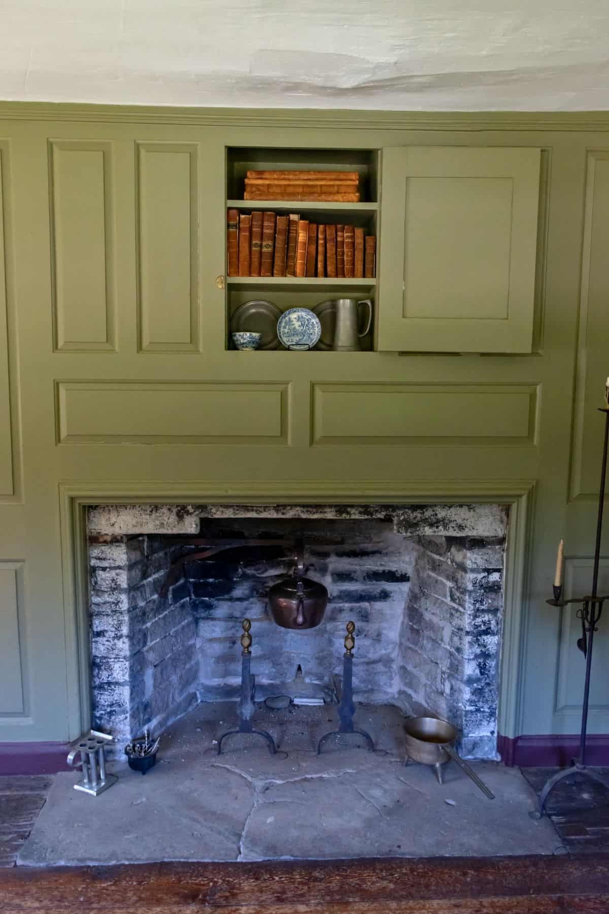 A fireplace with a sooty interior sits below a green built-in shelf holding books, a plate, a jug, and two small containers.