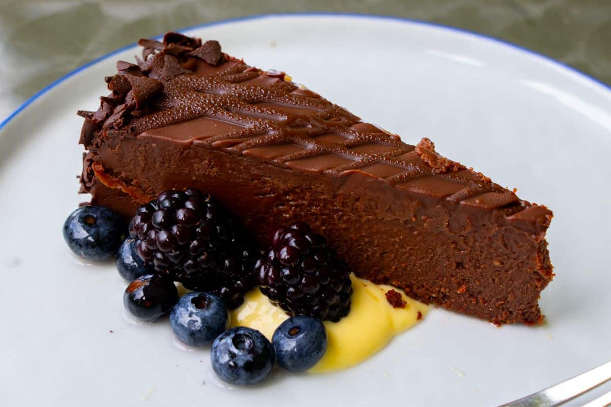 A slice of chocolate cheesecake on a white plate, garnished with blueberries, blackberries, and a dollop of yellow sauce.