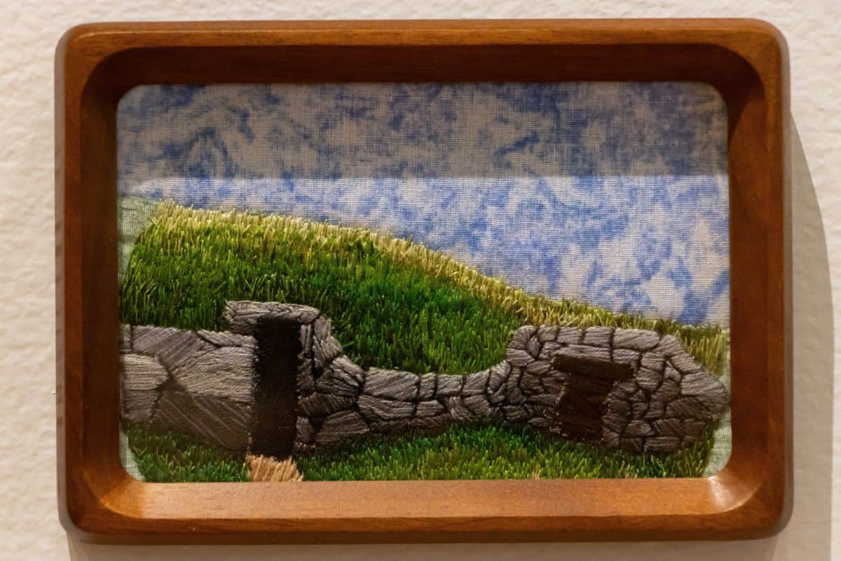 A framed embroidery depicts a grassy hill with two stone structures under a blue and white sky.