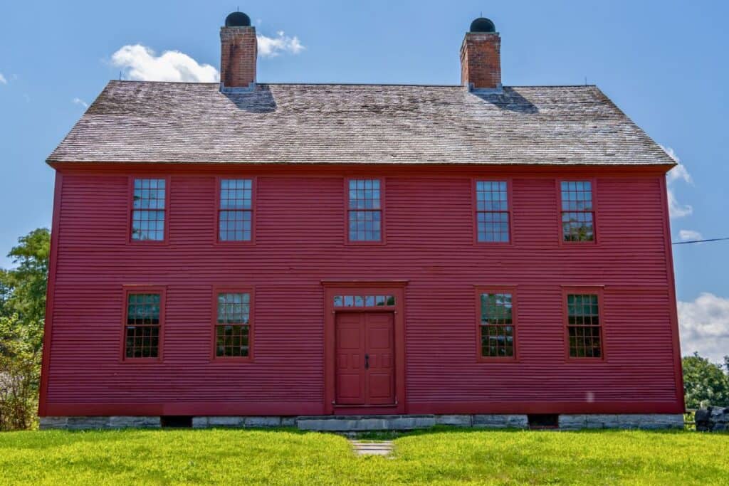A large red colonial-style house with two chimneys, symmetrical windows, and a central door, set against a blue sky and surrounded by grass.
