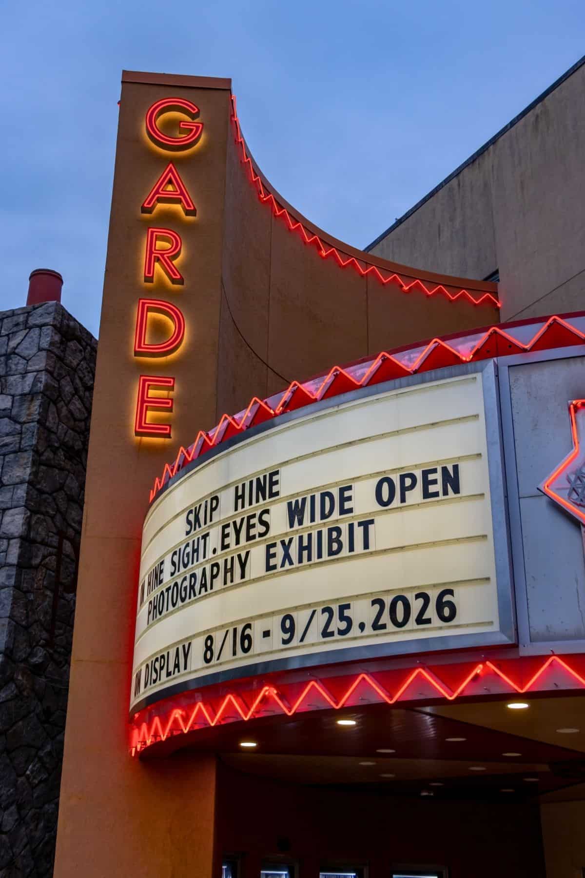 Theatre marquee displays “Skip Hine Fine Sight: Eyes Wide Open Photography Exhibit, on display 8/16 – 9/25/2026.” Neon “Garde” vertical sign is lit.