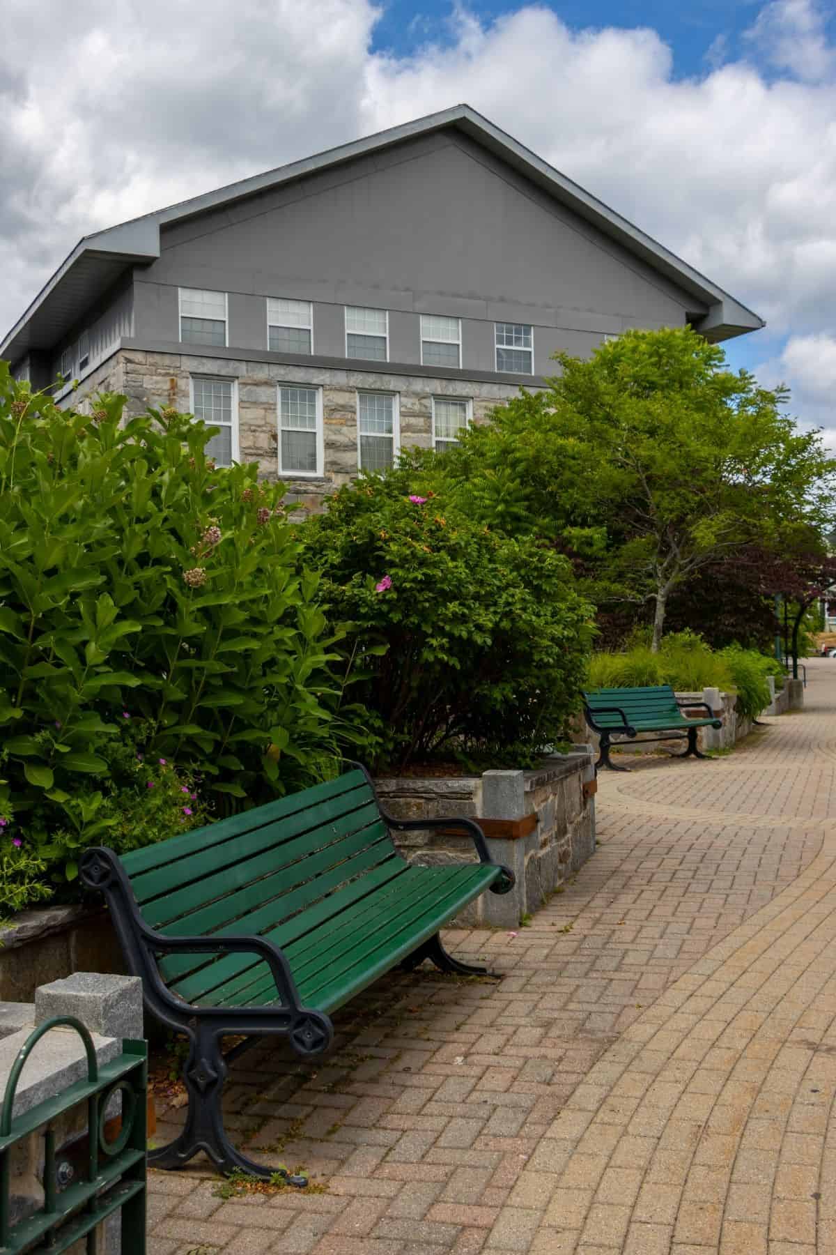 Green benches line a paved walkway beside bushes and trees, with a large gray building and partly cloudy sky in the background.