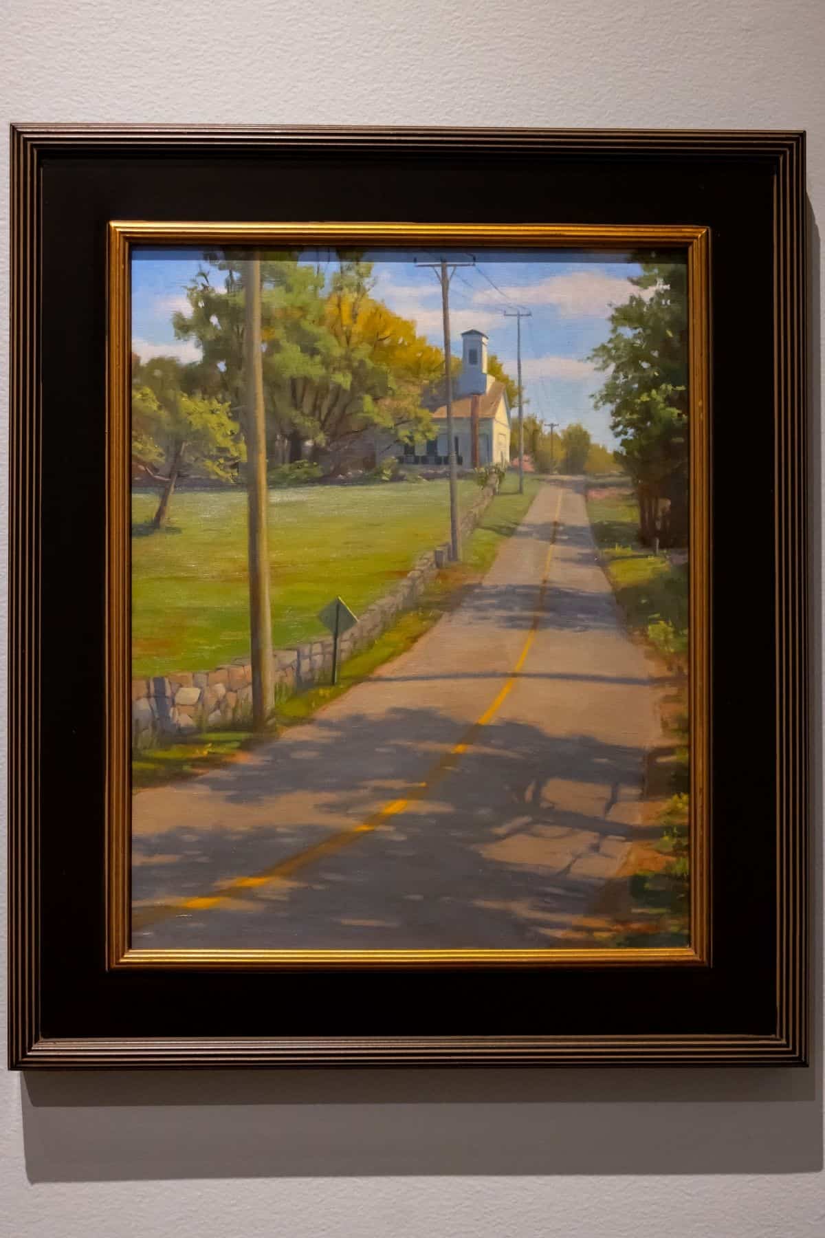 Framed painting of a rural road with a yellow line, stone wall, utility poles, green fields, trees, and a church or small building in the background under a partly cloudy sky.