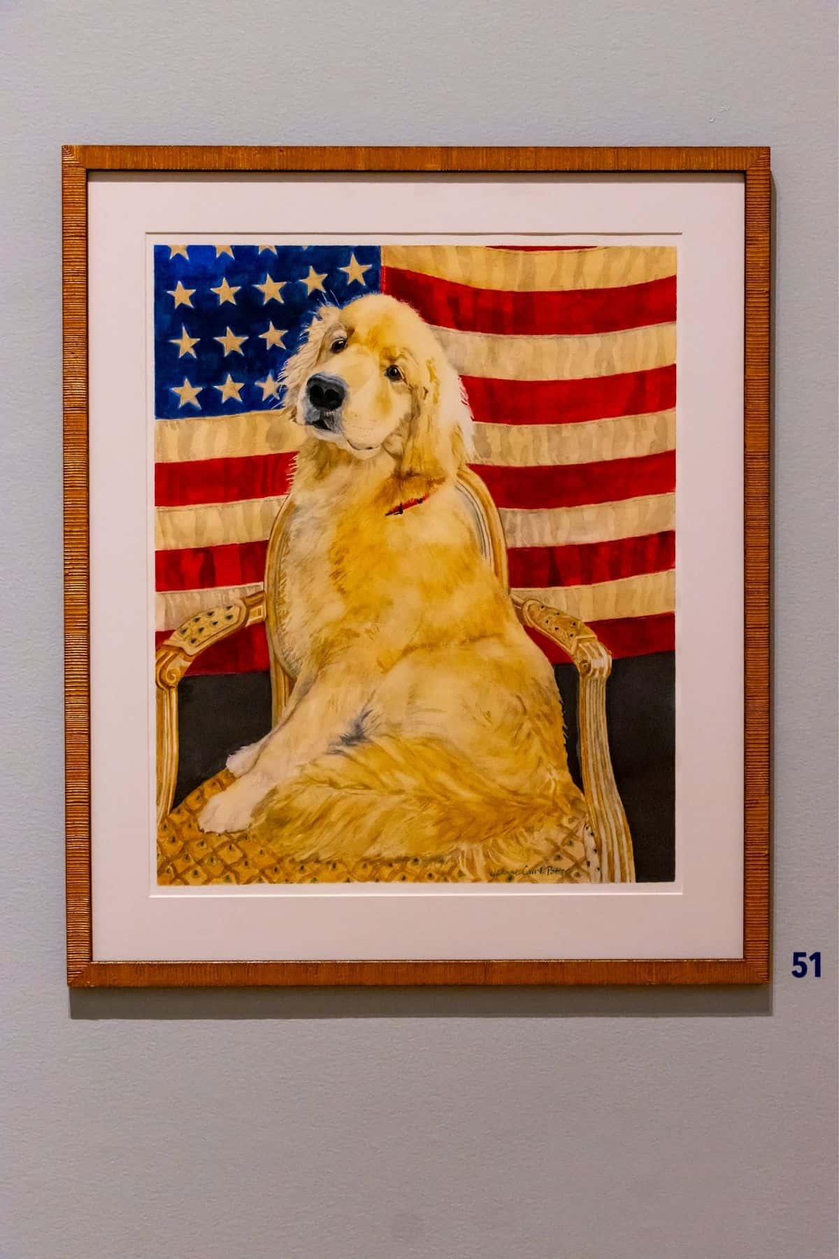 A framed painting of a golden retriever sitting on a chair in front of an American flag, displayed on a wall with a number 51 label.