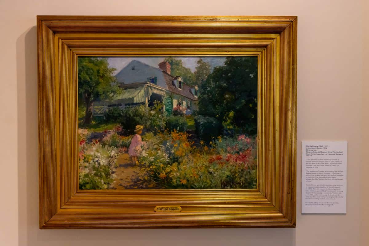 Framed painting of a garden scene with a person in a pink dress and hat among colorful flowers, with a house and trees in the background.