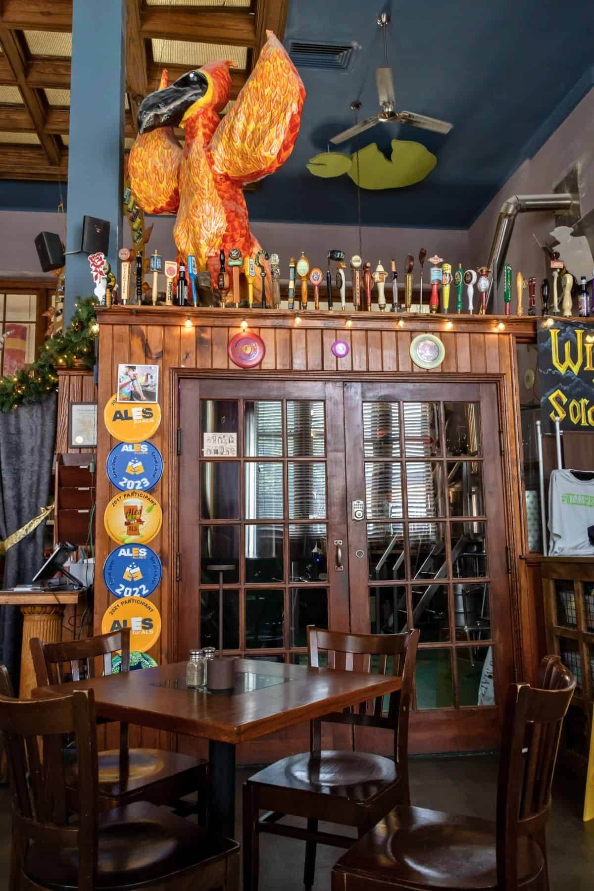 Wooden tables and chairs in front of double doors, with beer taps above and a large orange bird sculpture perched on top at a bar or pub.