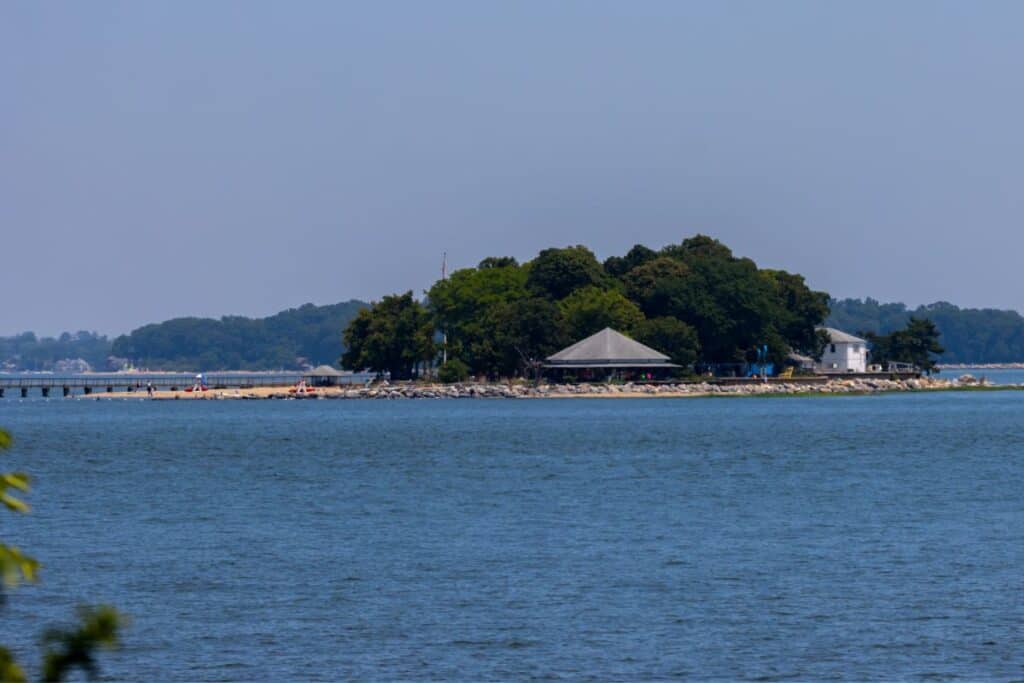 Small island with trees, a pavilion, and a white building, surrounded by water with a pier extending from the shore under a clear sky.