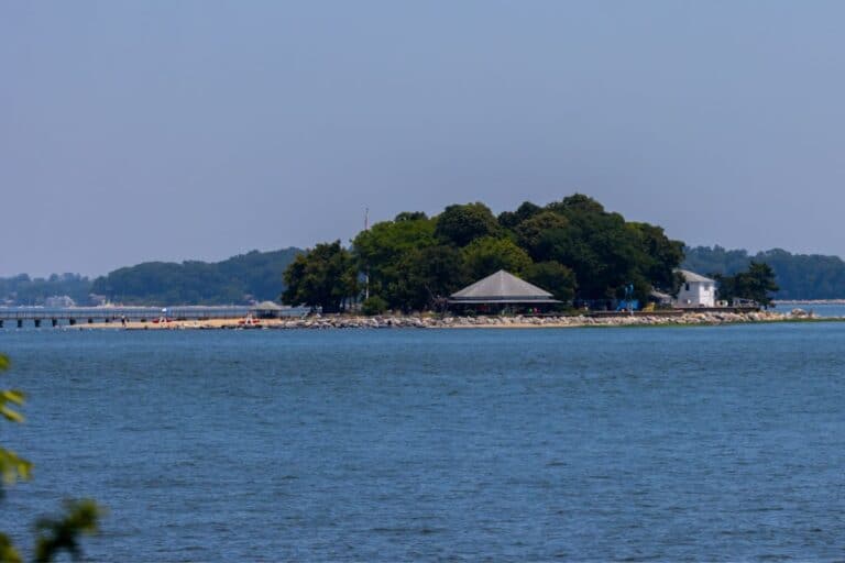Small island with trees, a pavilion, and a white building, surrounded by water with a pier extending from the shore under a clear sky.
