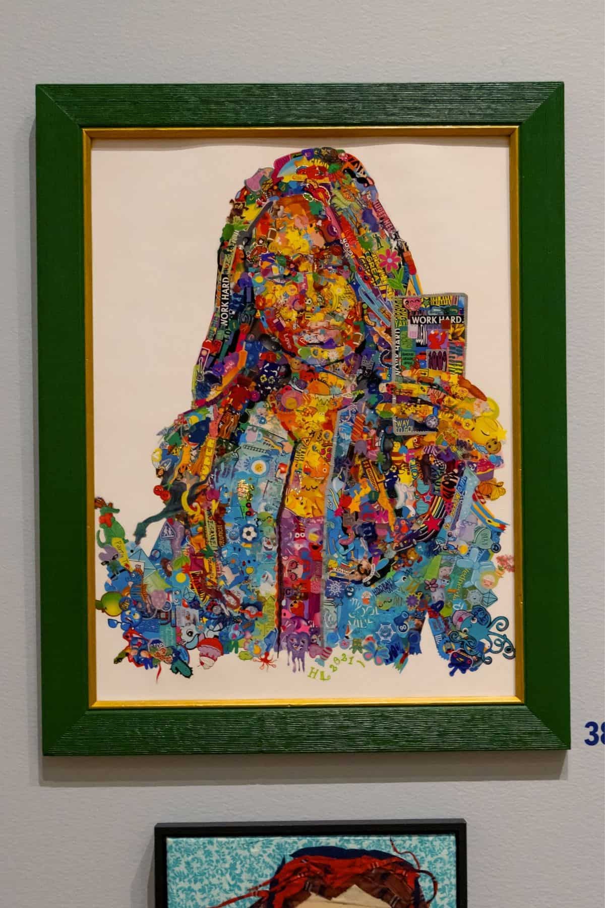 A colorful collage portrait of a woman holding a card, framed in green with a gold inner edge, displayed on a gray wall.