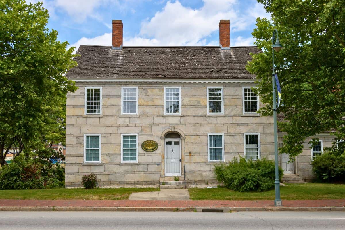 Two-story historic stone building with a symmetrical facade, white-framed windows, a central door, and two brick chimneys, surrounded by green trees and bushes along a sidewalk.