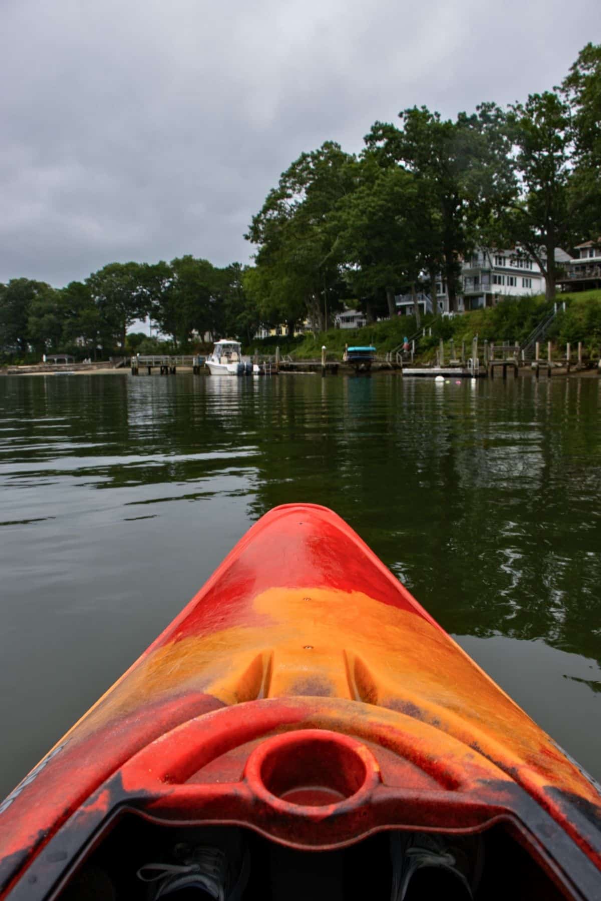 A view from a red-orange kayak on a calm lake, facing toward a shoreline with houses, docks, and boats under a cloudy sky.