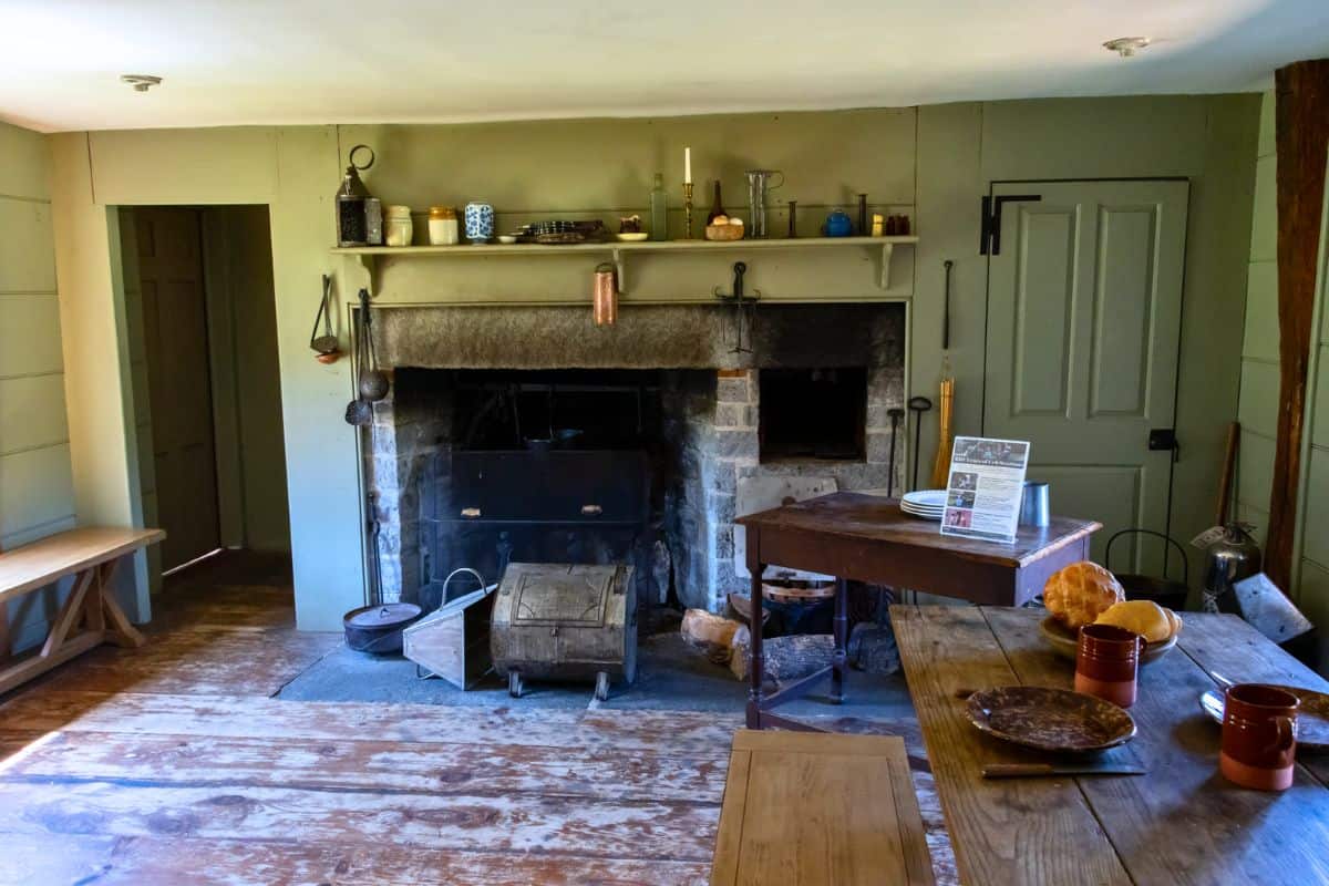 A rustic kitchen with a large stone fireplace, wooden table and benches, cookware on shelves, and various household items arranged neatly.