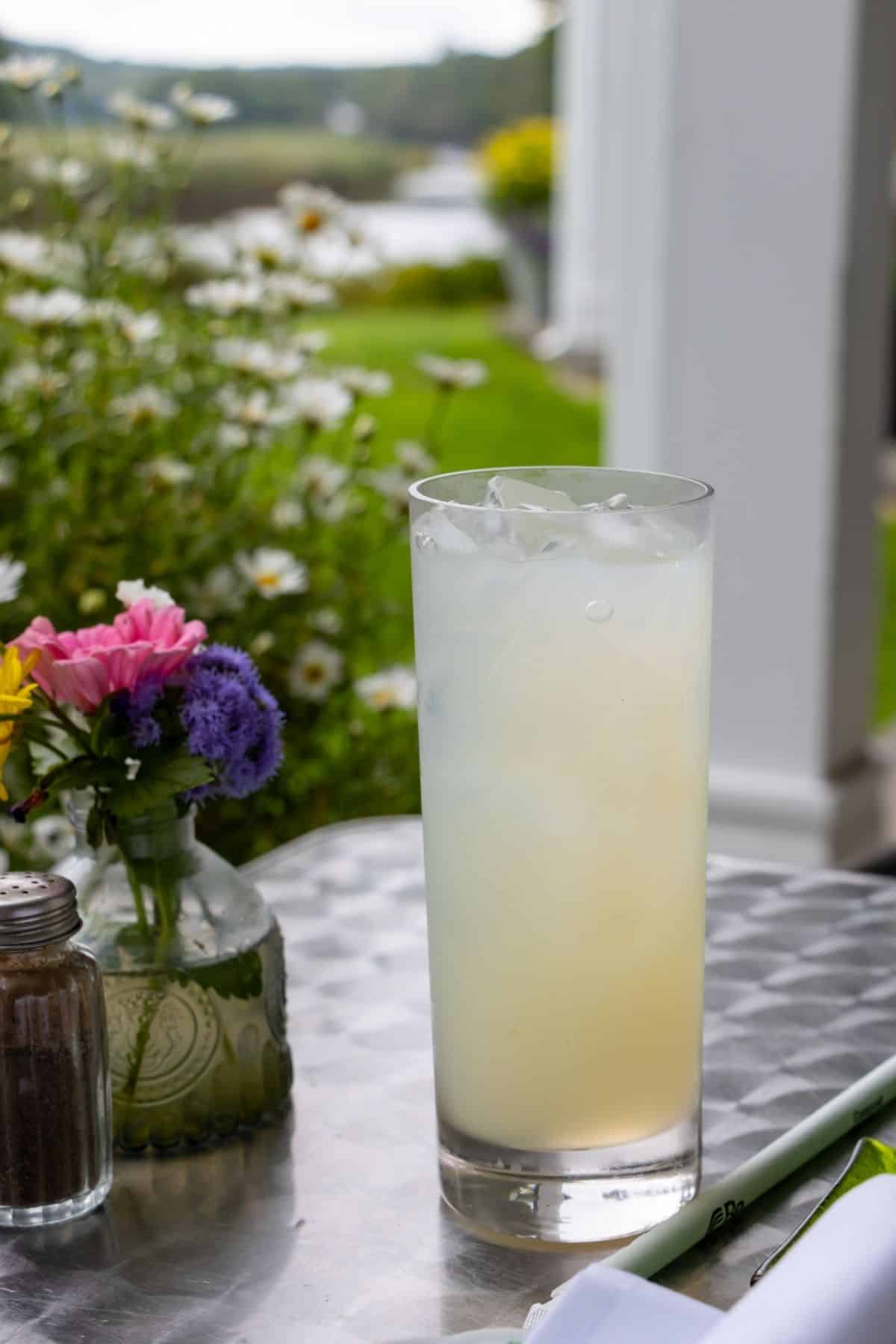 A tall glass of iced lemonade sits on an outdoor table next to a small vase of flowers and a pepper shaker, with greenery and flowers in the background.