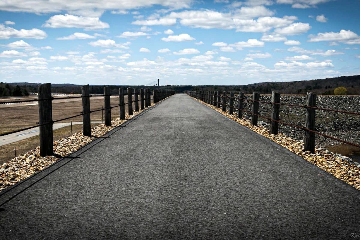 A paved path bordered by rocks and wooden railings extends straight into the distance under a partly cloudy sky.