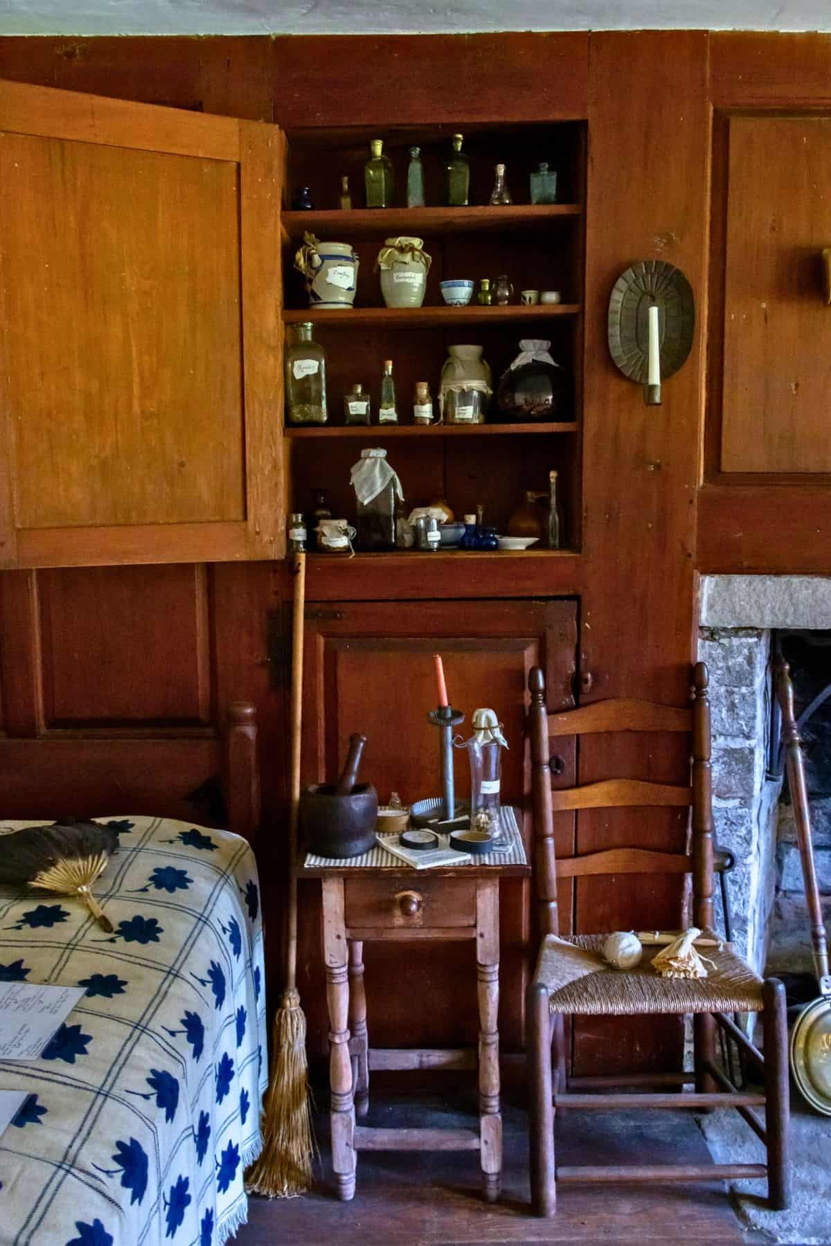 Wooden table with a candle, mortar and pestle, and broom beside a ladder-back chair; shelves with jars and crockery on a wooden wall near a bed and stone fireplace.