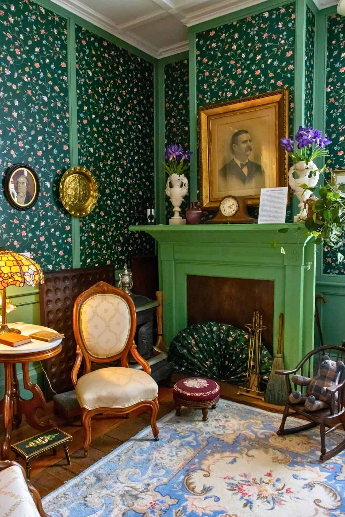 Victorian-style living room with green floral wallpaper, a green fireplace, framed portraits, antique furniture, and various decorative items including a brass lamp and purple flowers.
