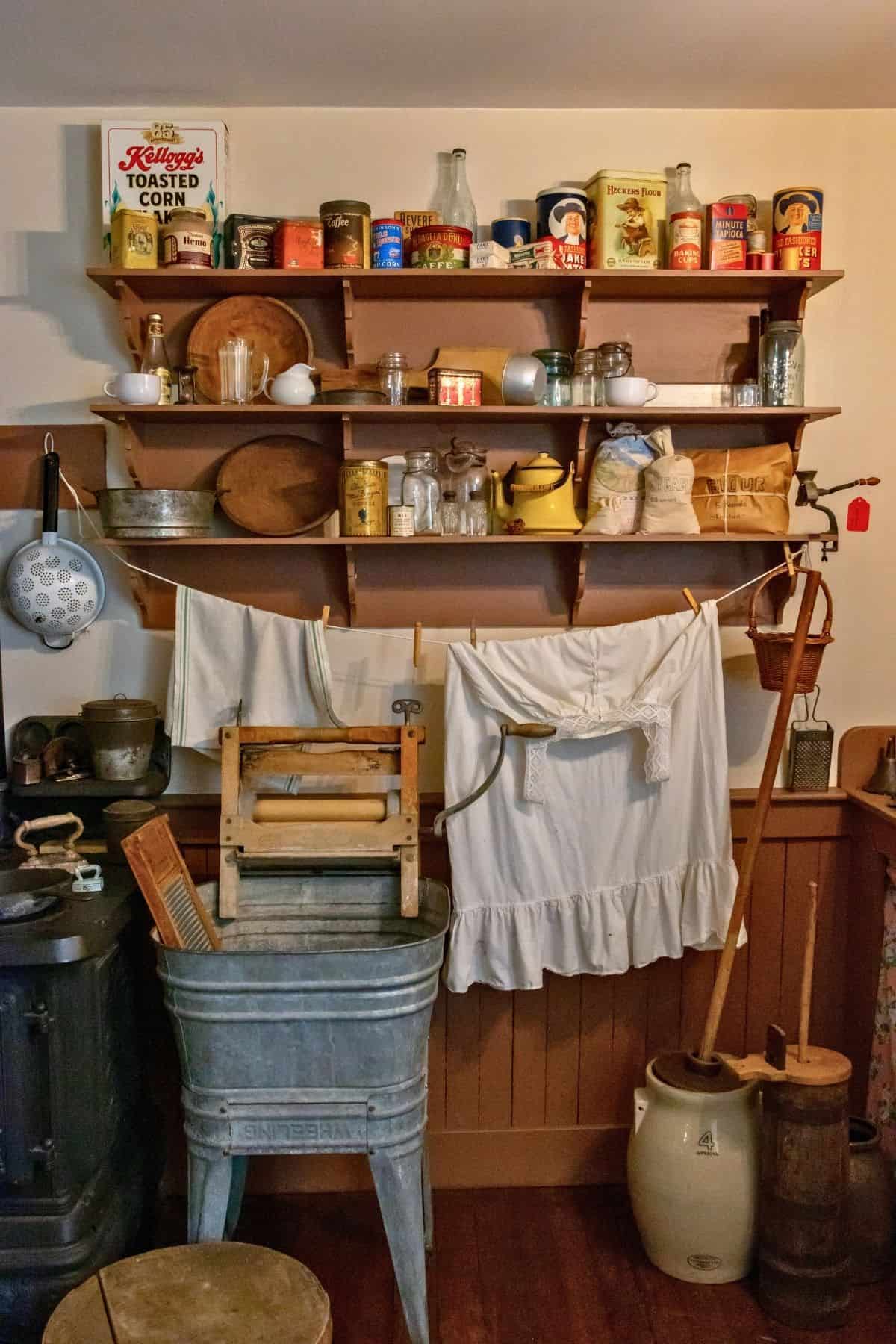 Vintage kitchen scene with shelves of canned goods, cookware, and teapots above a washboard, wringer, drying linens, and household tools, recreating an early 1900s domestic setting.