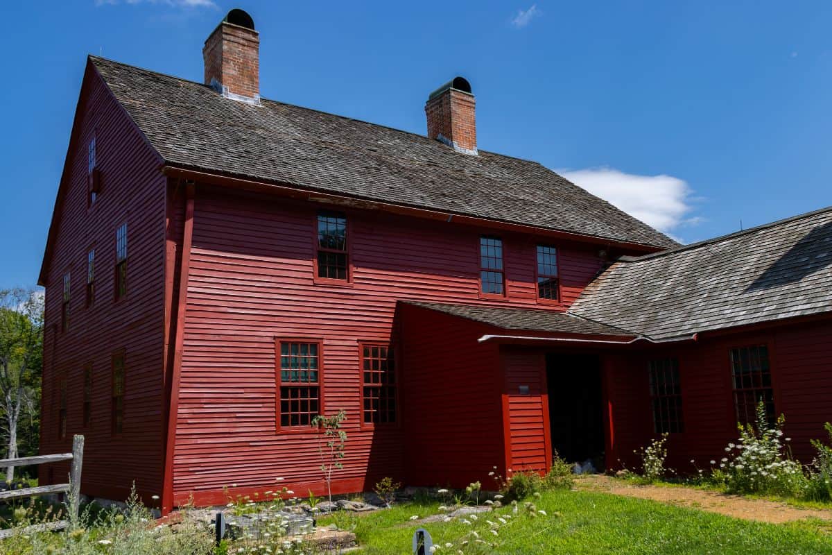 Large red wooden building with multiple chimneys and windows, set against a blue sky with some clouds. Grassy area and wildflowers are in the foreground.
