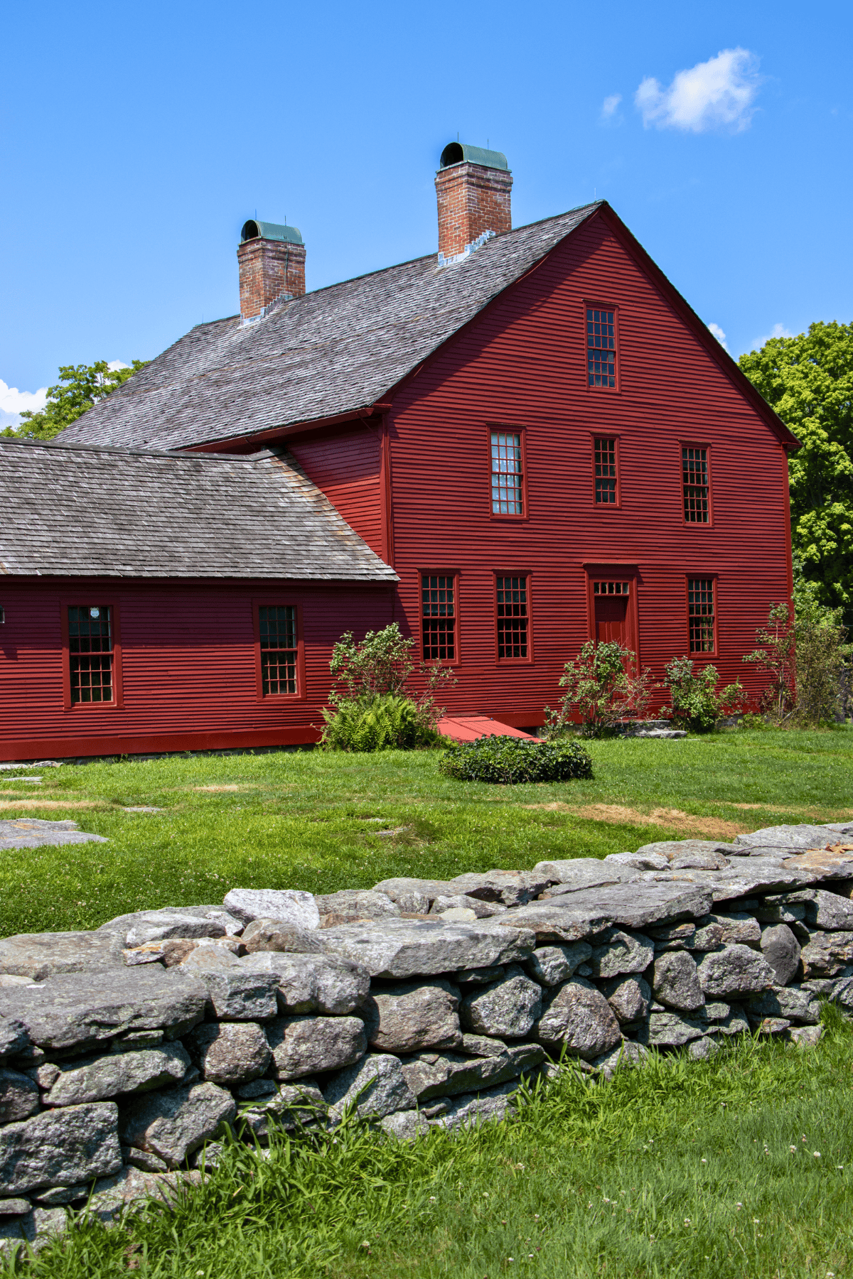 A large red wooden house with two chimneys stands behind a stone wall and a grassy lawn under a blue sky with a few clouds.