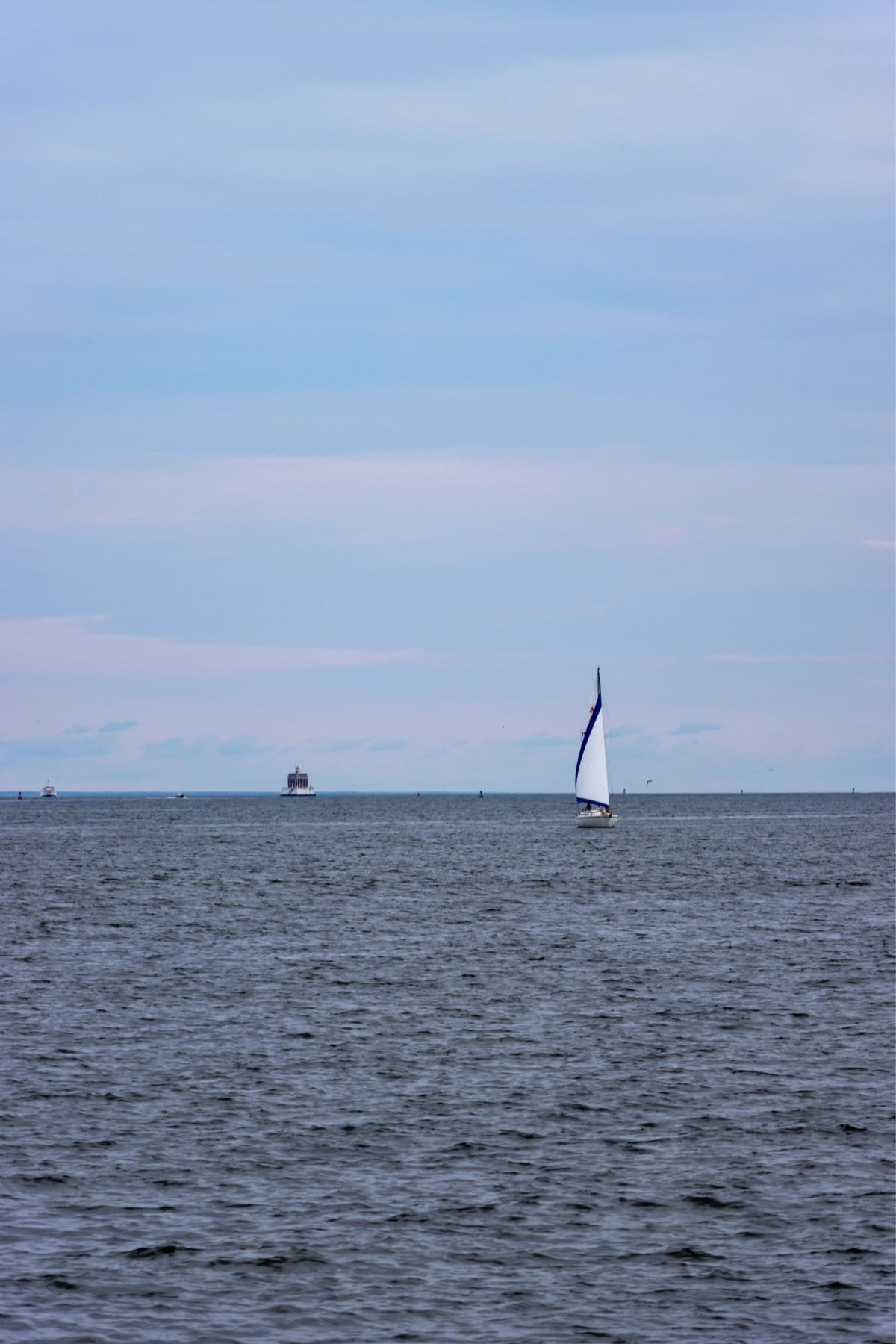 A sailboat with a white and blue sail moves on calm water under a cloudy sky, with a cargo ship visible in the distant background.