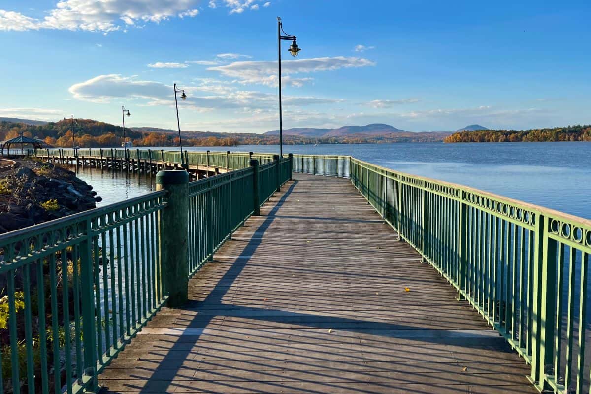A wooden boardwalk with green railings extends over a lake, with hills and trees in the background under a blue sky with clouds.