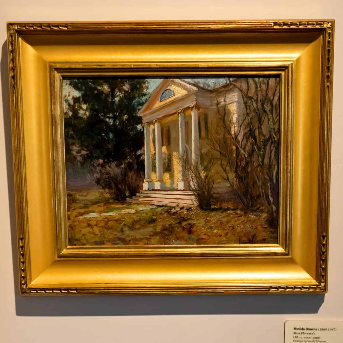 A framed painting depicts a small classical building with columns, surrounded by trees and bushes, under a cloudy sky.