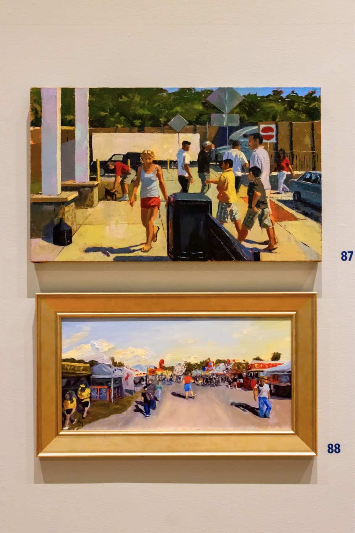 Two framed paintings hang on a wall, each depicting outdoor scenes with people. The upper painting is numbered 87, and the lower painting is numbered 88.