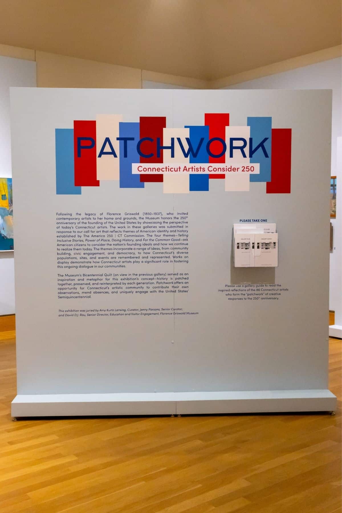 Exhibit display panel titled “PATCHWORK: Connecticut Artists Consider 250” with colored blocks and informational text, set on a wooden floor in an art gallery.