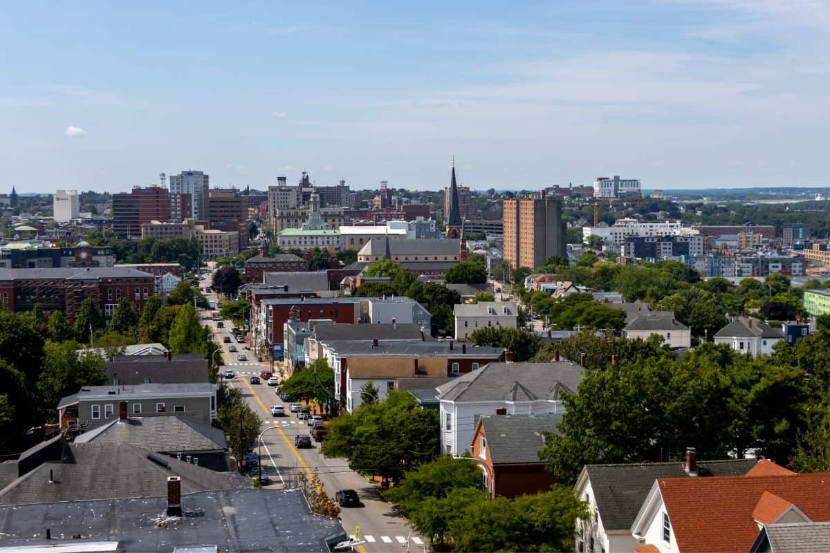 A cityscape showing residential homes in the foreground and taller buildings in the background under a partly cloudy sky. Trees are scattered throughout the area.