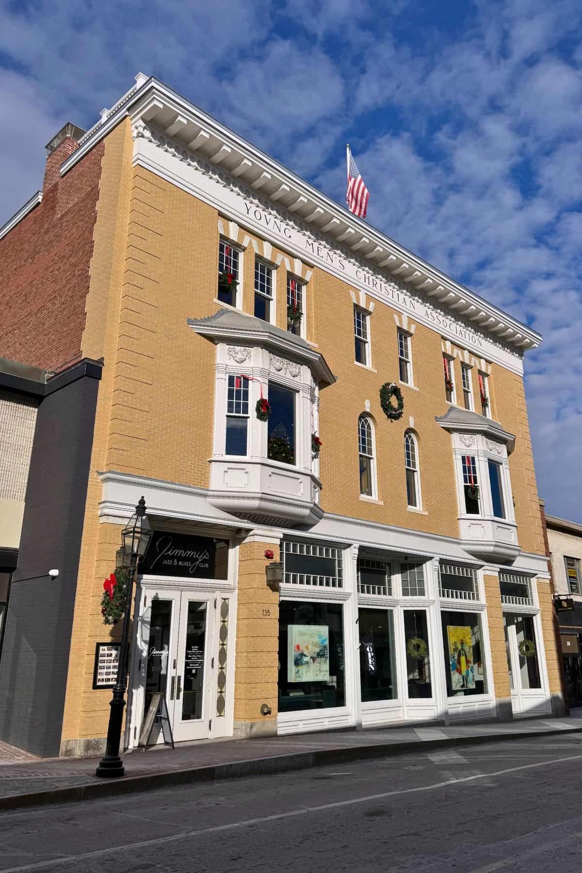 Three-story brick building with “Young Men’s Christian Association” on the front, U.S. flag on the roof, and storefronts at street level, decorated with wreaths and holiday garlands.