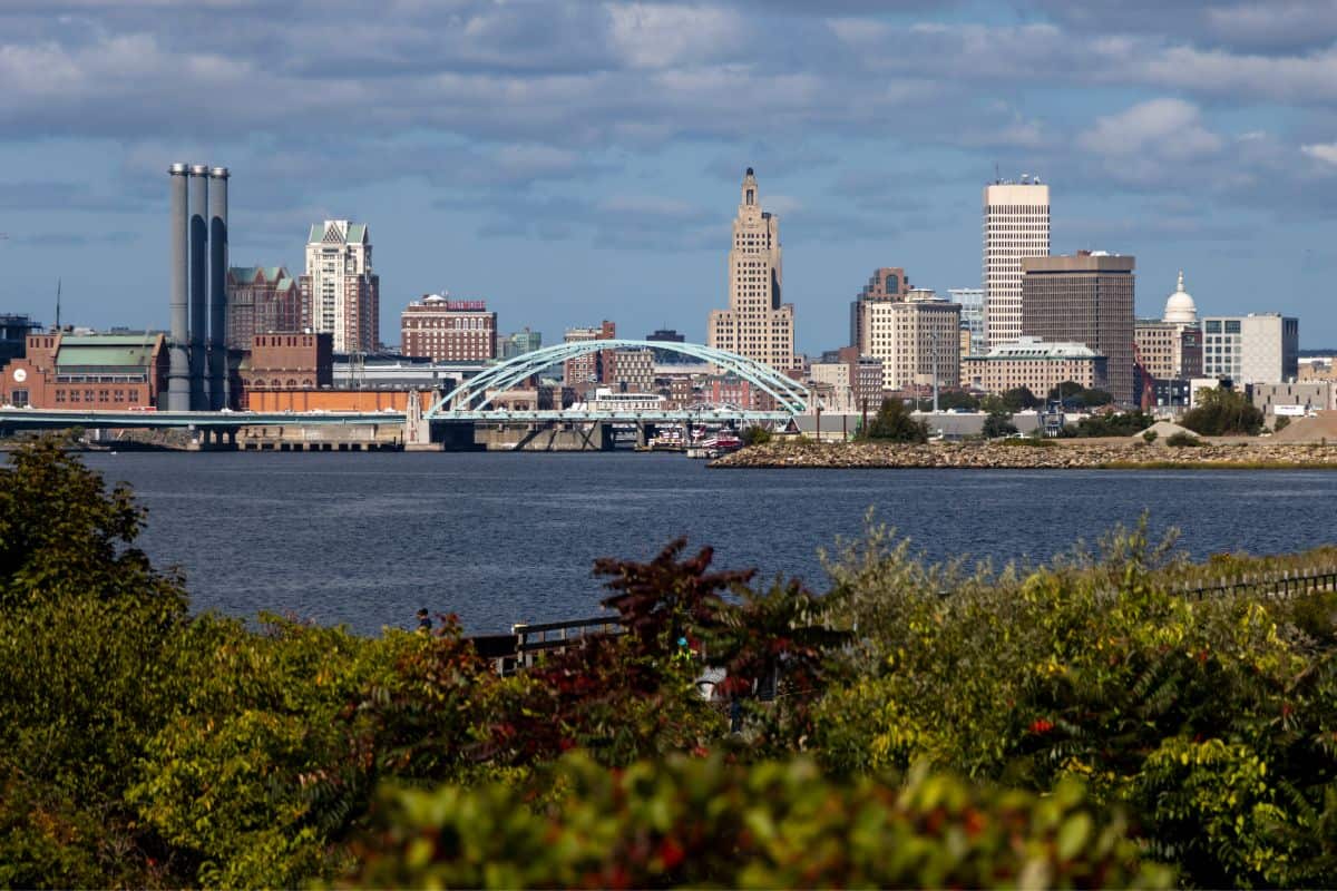 Providence city skyline with prominent buildings and a bridge, viewed across a body of water with greenery in the foreground under a partly cloudy sky.