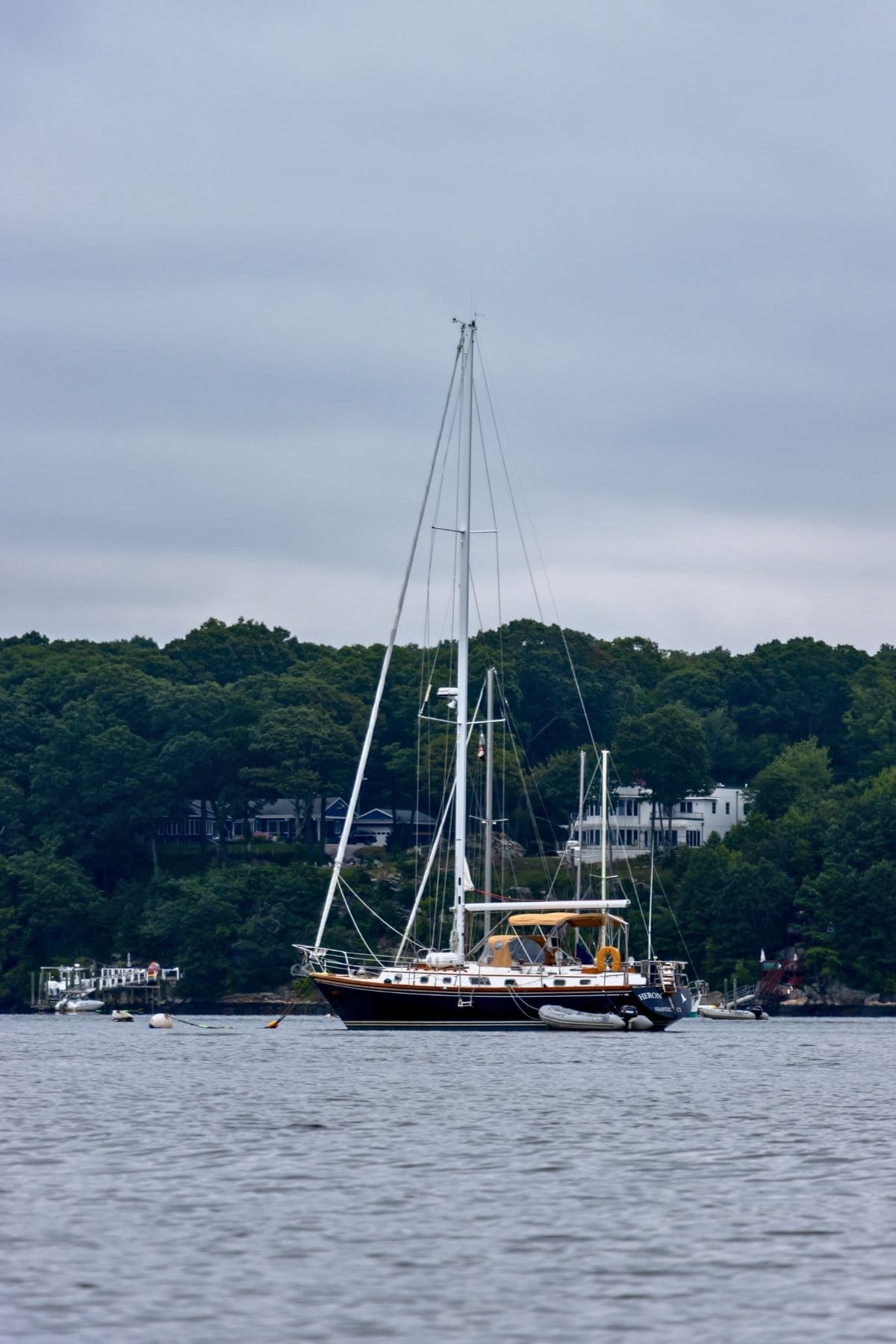 A sailboat is anchored on calm water near a tree-lined shoreline with several houses visible in the background under a cloudy sky.