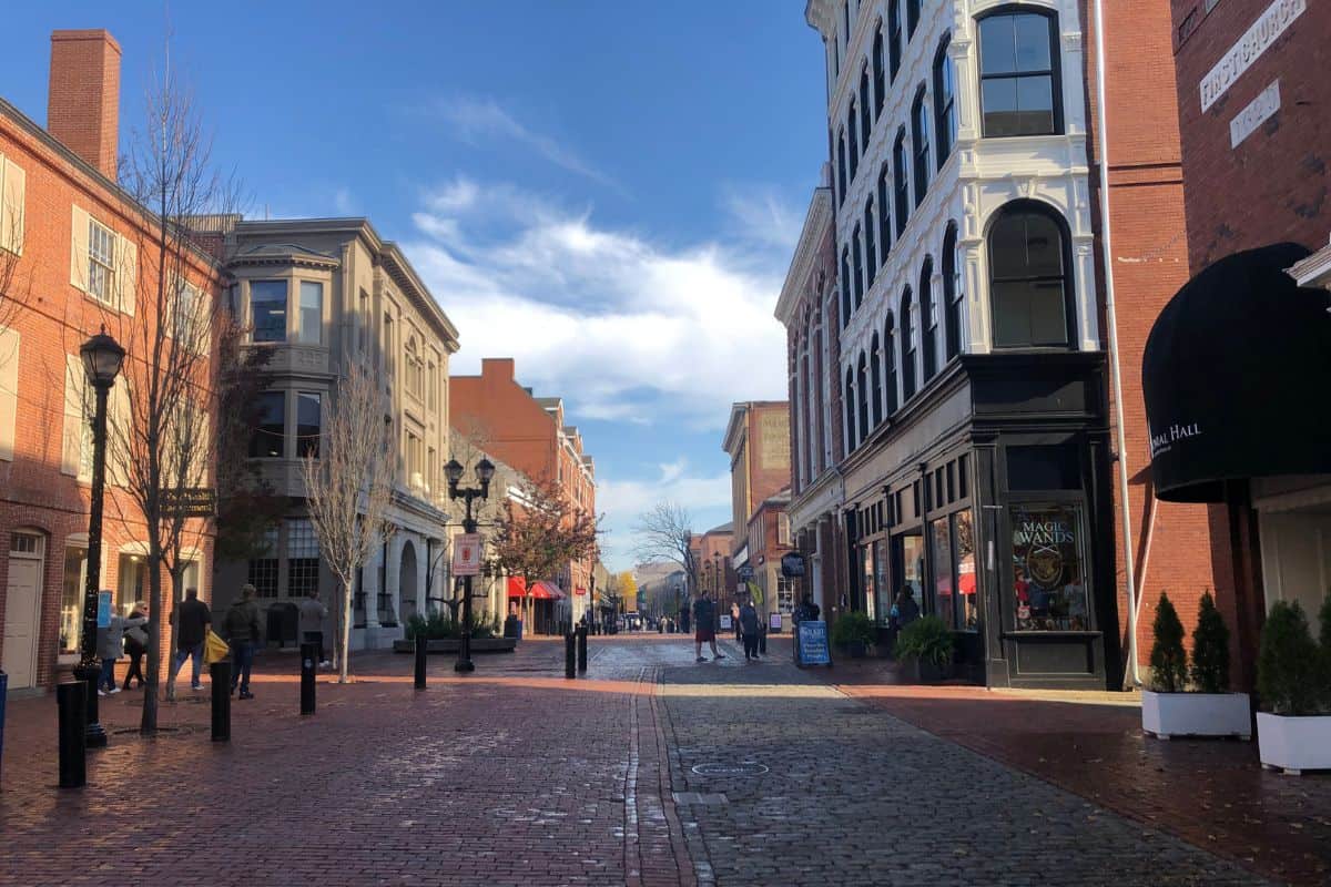 Brick-paved street lined with historic buildings, shops, and pedestrians under a partly cloudy sky.