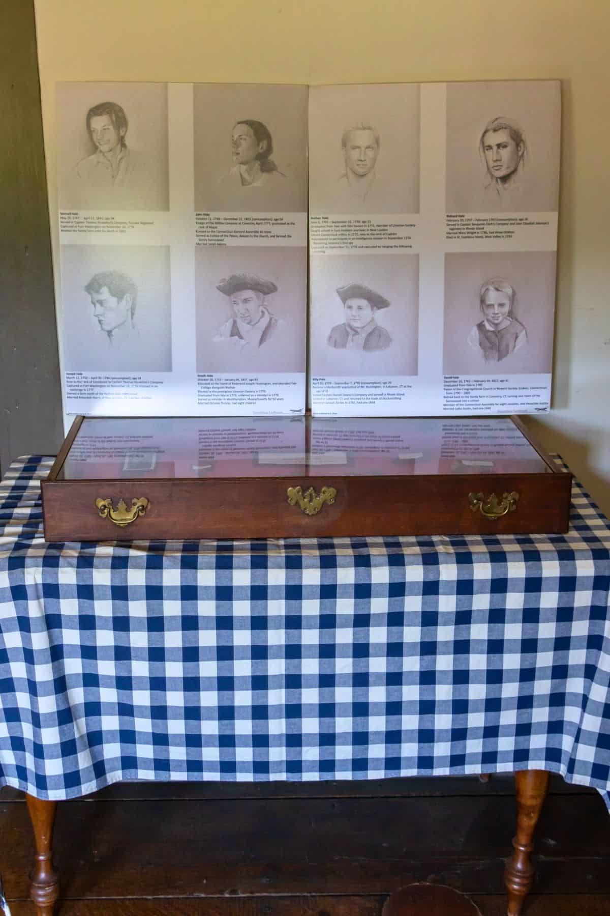 A display case on a blue and white checkered table holds documents; behind it are drawings and descriptions of eight historical figures mounted on panels.