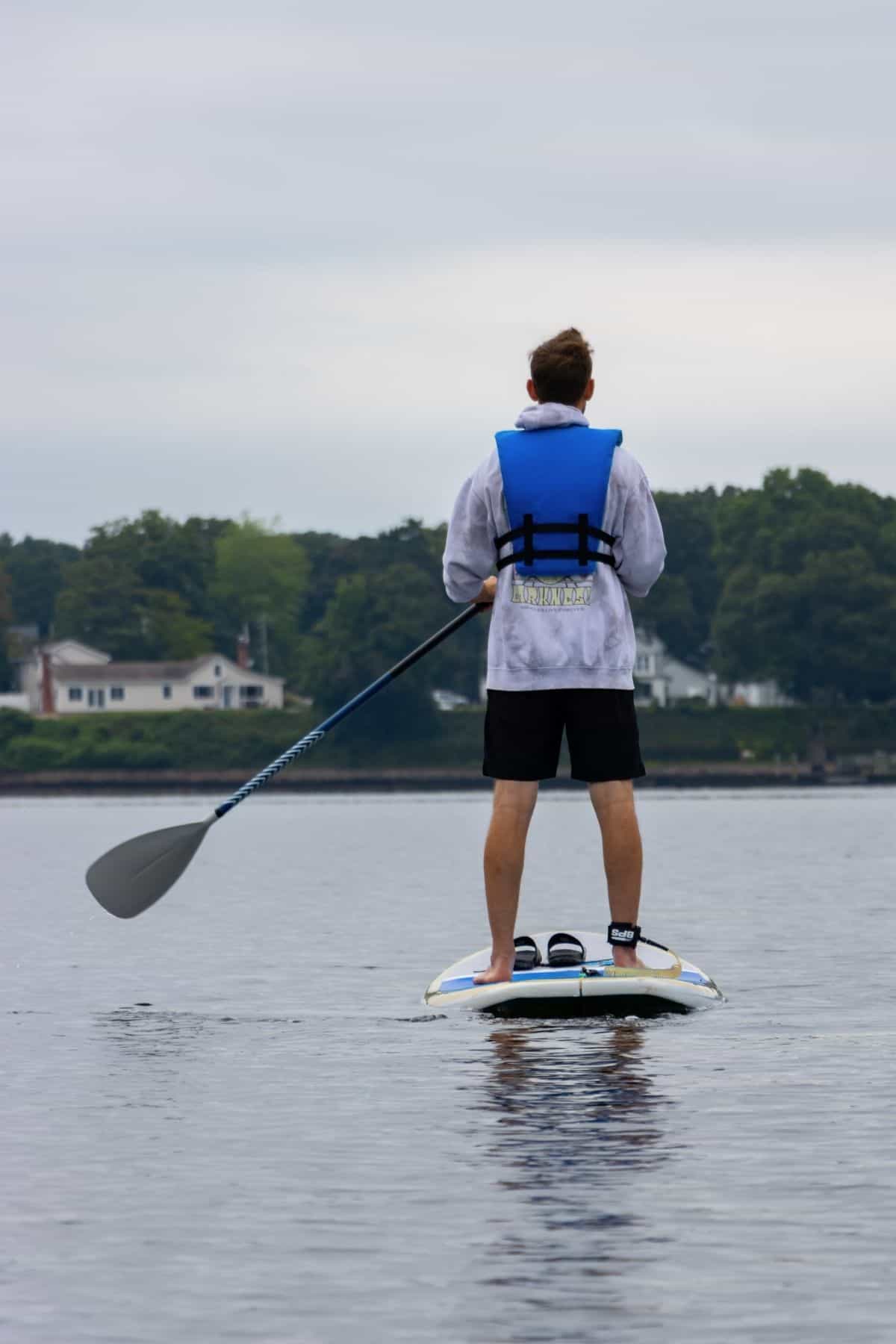 A person wearing a blue life jacket stands on a paddleboard, holding a paddle, on calm water near a shoreline with houses and trees.