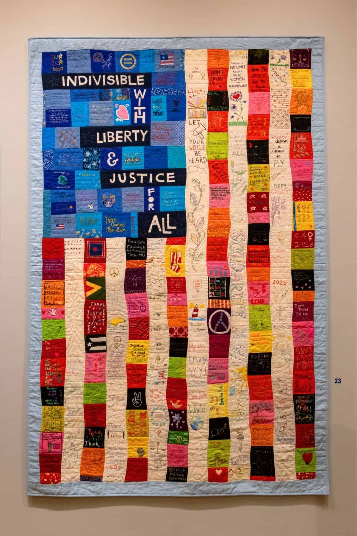 A handmade quilt designed like the American flag with colorful squares, handwritten messages, and the words “INDIVISIBLE WITH LIBERTY & JUSTICE FOR ALL” in the blue section.