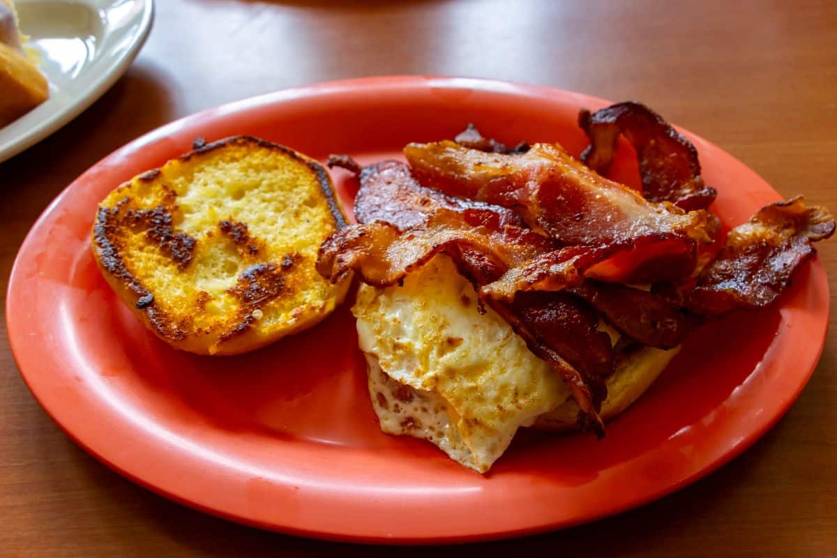 A breakfast sandwich with bacon, a fried egg, and a toasted bun is served on an orange plate on a wooden table.
