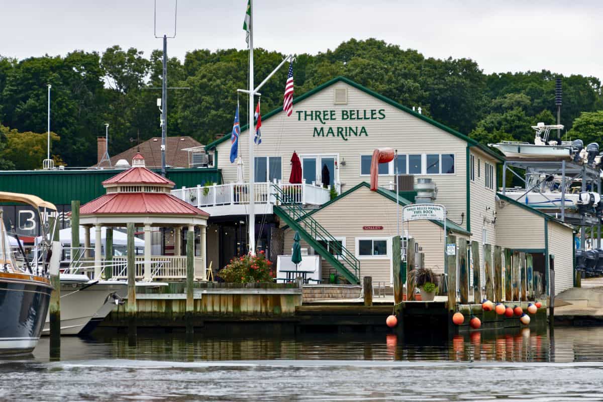 A marina building labeled “Three Belles Marina” stands by the water, with boats docked nearby and a gazebo, flags, and trees in the background.