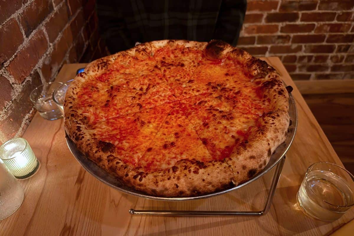 A large cheese pizza on a metal stand sits on a wooden table with a glass of water, a candle, and brick walls in the background.