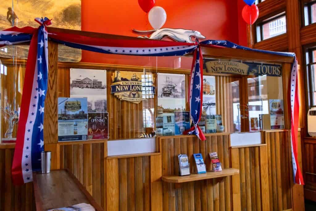 Wooden ticket counter decorated with red, white, and blue star banners, historical posters, brochures, and balloons, inside a brightly lit visitor center.
