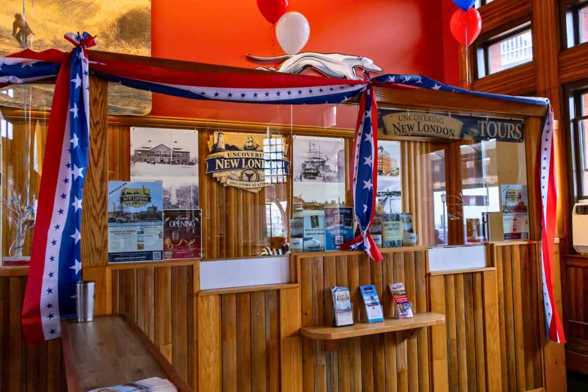 Wooden ticket counter decorated with red, white, and blue star banners, historical posters, brochures, and balloons, inside a brightly lit visitor center.