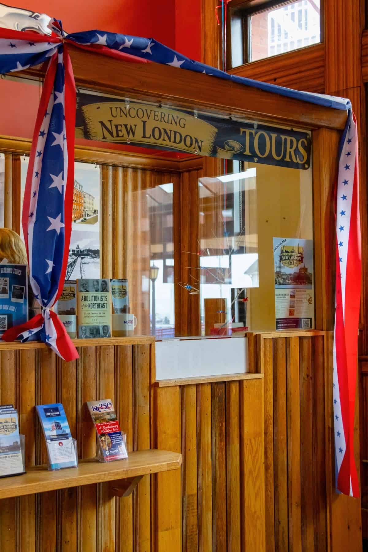 Wooden tour information booth decorated with red, white, and blue star ribbons, displaying brochures and a sign reading “UNCOVERING NEW LONDON TOURS.”.