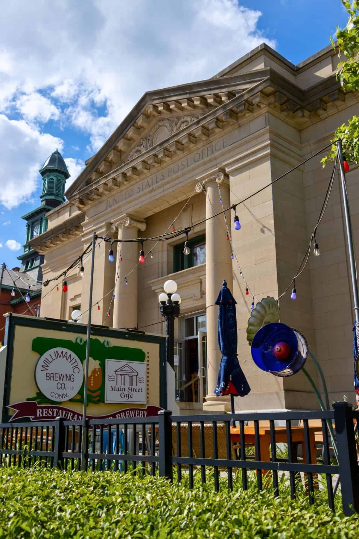 A historic post office building with columns, string lights, and signs for Willimantic Brewing Co. and Main Street Cafe in the foreground. Blue sky with clouds overhead.